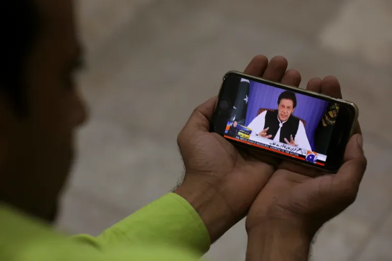 A journalist poses with a cell phone displaying a speech by Imran Khan